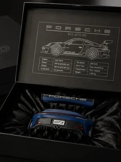 Dois porta-chaves do Porsche GT3 RS em azul: um acomodado no forro de cetim da caixa aberta, sob a placa gravada na tampa, e outro sobre a mesa ao lado da caixa fechada